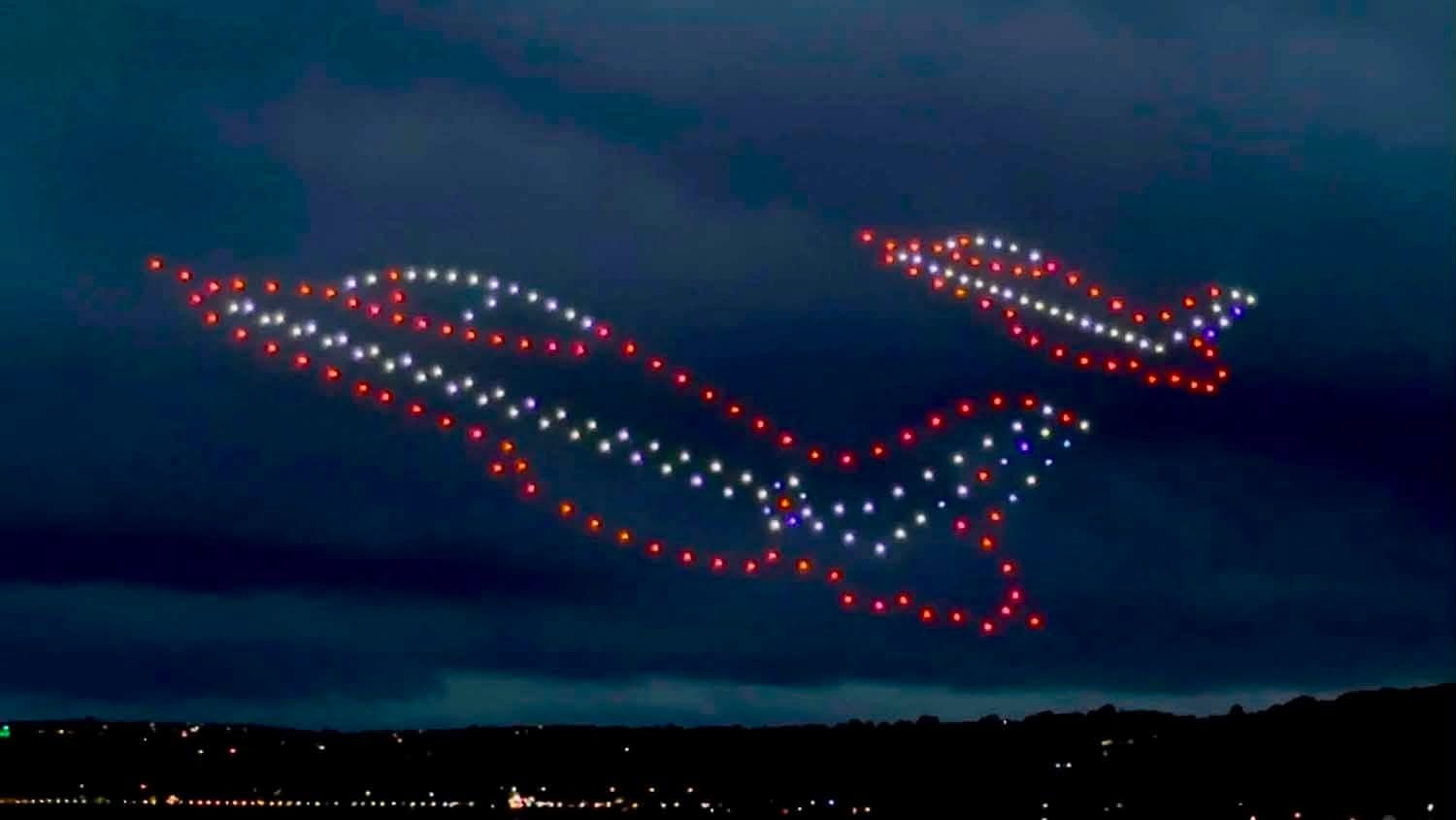 A drone formation of aircraft in red, white and blue flying in formation across a dark sky