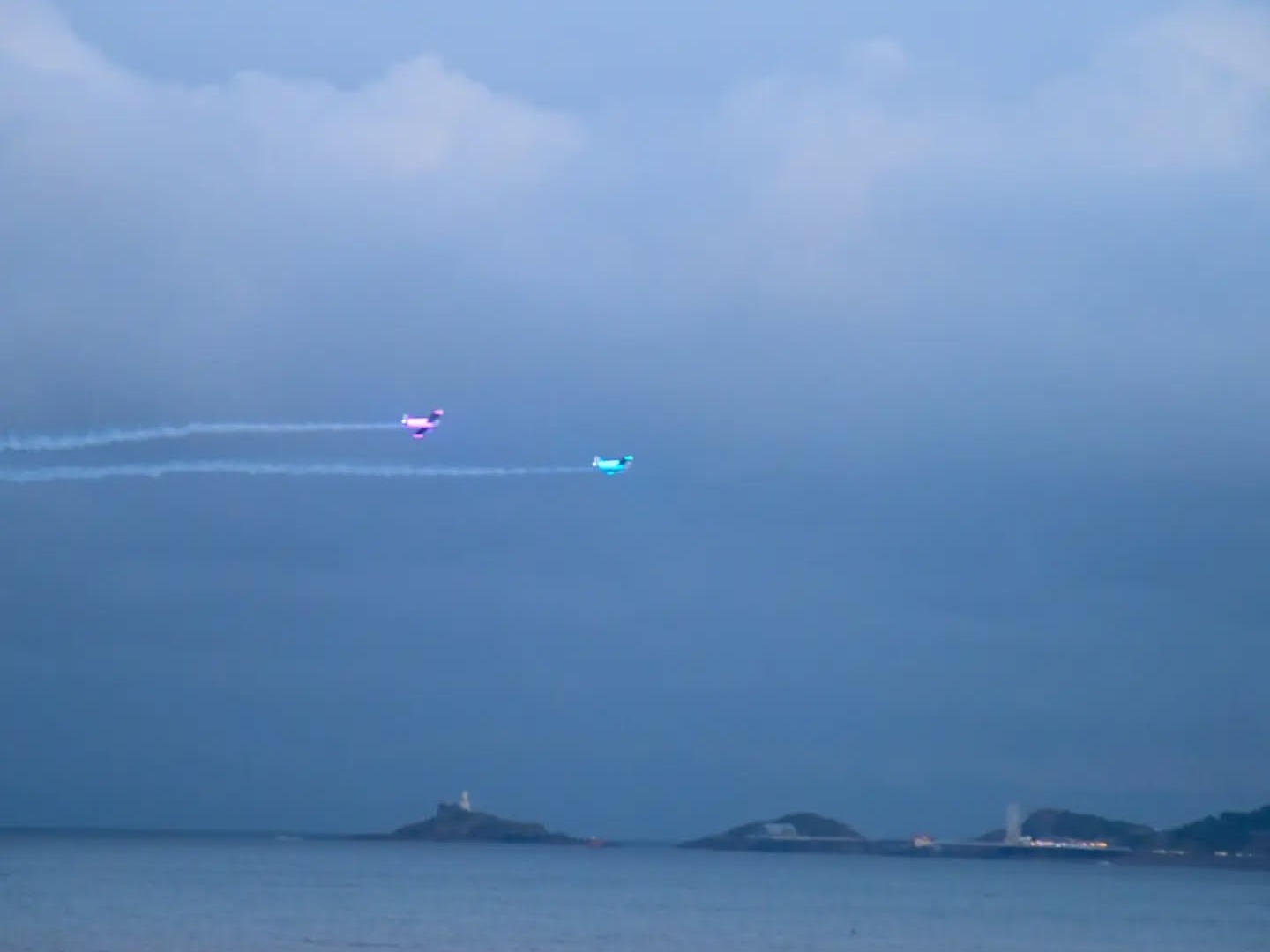 Two Firebirds display aircraft lit in pink and blue flying low over Swansea Bay at dusk with Mumbles lighthouse behind