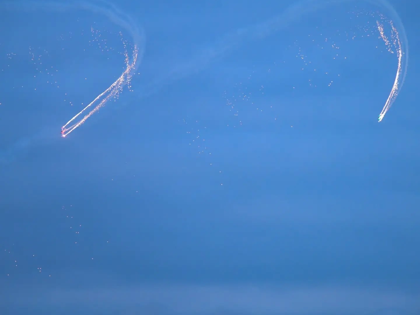 Display aircraft arcing across a dusk sky trailing bright pyrotechnic sparks over Swansea Bay