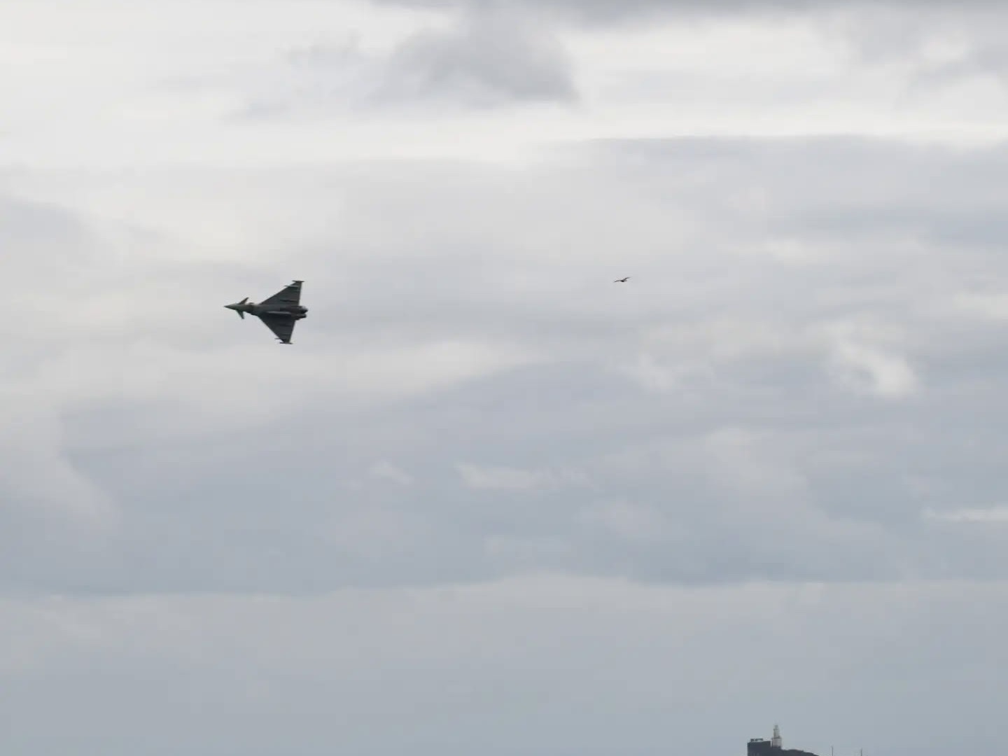 An RAF Typhoon flying over Swansea Bay with Mumbles lighthouse on the headland below and small boats moored offshore