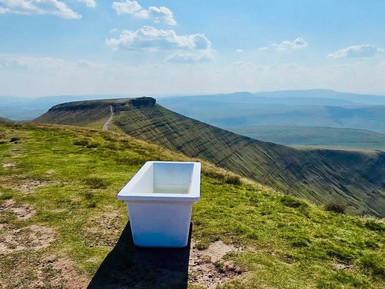 A white bathtub standing alone on the grassy summit ridge of Pen y Fan, the flat-topped peak of Corn Du behind under a blue sky.