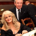 Bonnie Tyler seated at a table signing a large ceremonial book, with her husband Robert Sullivan standing behind her