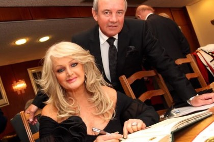 Bonnie Tyler seated at a table signing a large ceremonial book, with her husband Robert Sullivan standing behind her