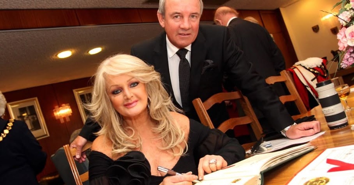 Bonnie Tyler seated at a table signing a large ceremonial book, with her husband Robert Sullivan standing behind her