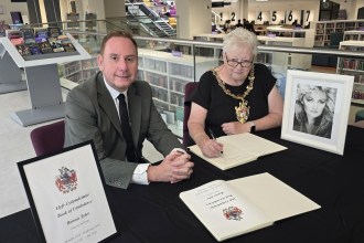 Cllr Rob Stewart and Lord Mayor Cllr Penny Matthews sitting at a table signing the Bonnie Tyler book of condolence inside Y Storfa library