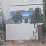 A man on a ladder rolls white paint over a large wall mural, with a painted banner, trees and Brunel's black top hat still visible beneath.