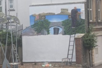 A man on a ladder rolls white paint over a large wall mural, with a painted banner, trees and Brunel's black top hat still visible beneath.