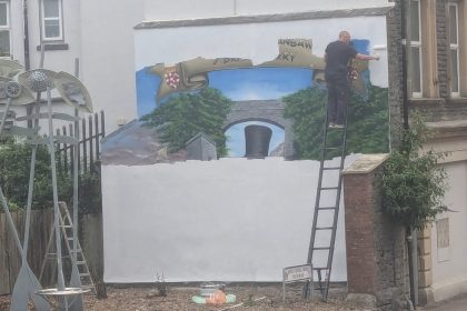 A man on a ladder rolls white paint over a large wall mural, with a painted banner, trees and Brunel's black top hat still visible beneath.