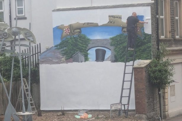 A man on a ladder rolls white paint over a large wall mural, with a painted banner, trees and Brunel's black top hat still visible beneath.