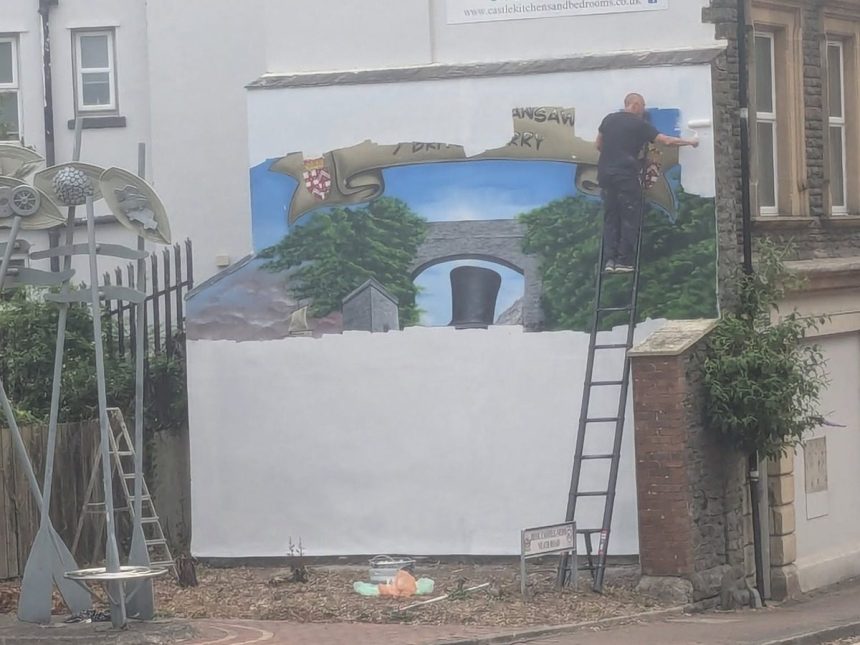 A man on a ladder rolls white paint over a large wall mural, with a painted banner, trees and Brunel's black top hat still visible beneath.