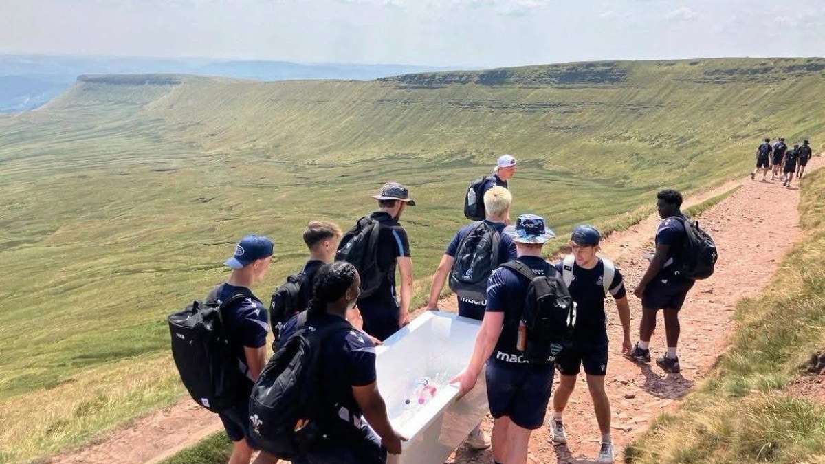Young men in navy Cardiff Rugby training kit carry a white bathtub down a stone path, the escarpment stretching away behind.