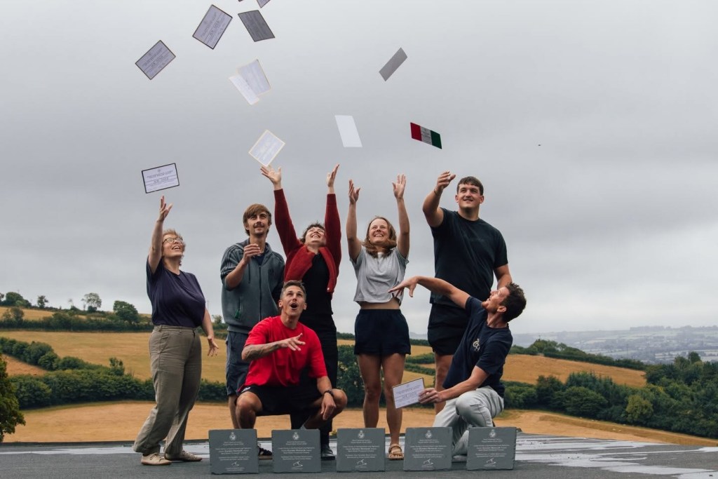 Seven members of the Caws Teifi team throwing award certificates into the air on a hilltop, with slate trophies lined up in front of them