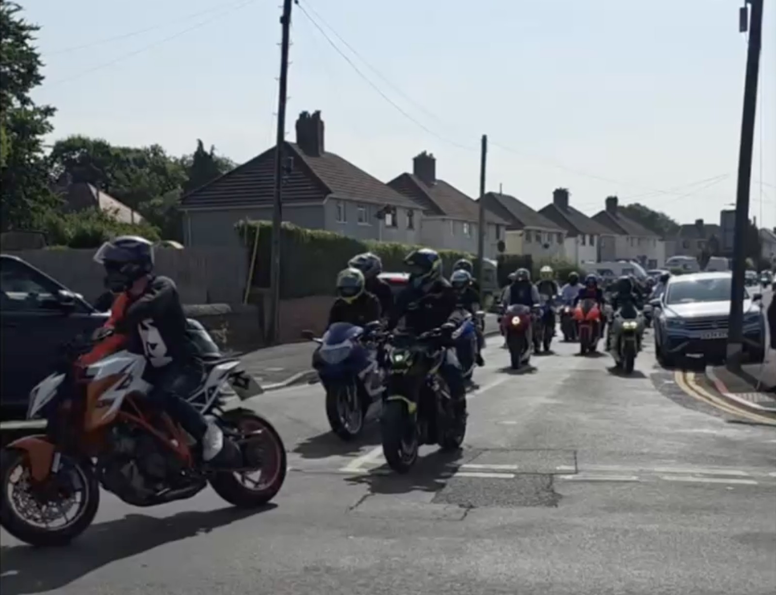 Motorbikes riding along a residential street with people watching from the roadside