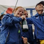 Lou Boorman, with red hair and in blue sailing jacket, laughing as she holds a champagne bottle among her celebrating crew on the yacht's deck