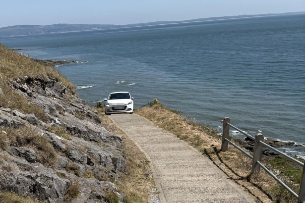 A white hatchback part-way along a narrow clifftop coastal footpath above the rocks, with the sea beyond.