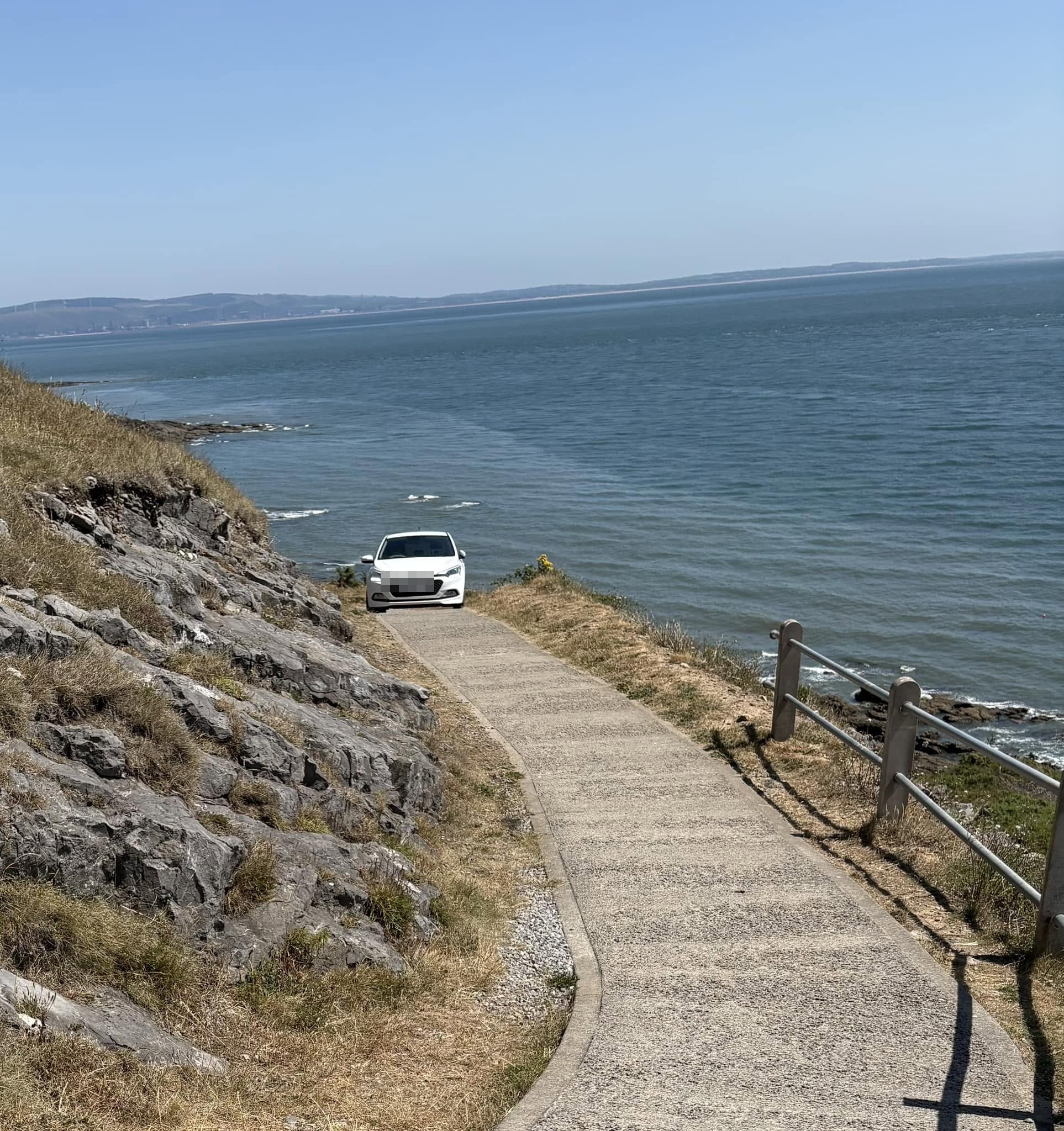 A white hatchback part-way along a narrow clifftop coastal footpath above the rocks, with the sea beyond.