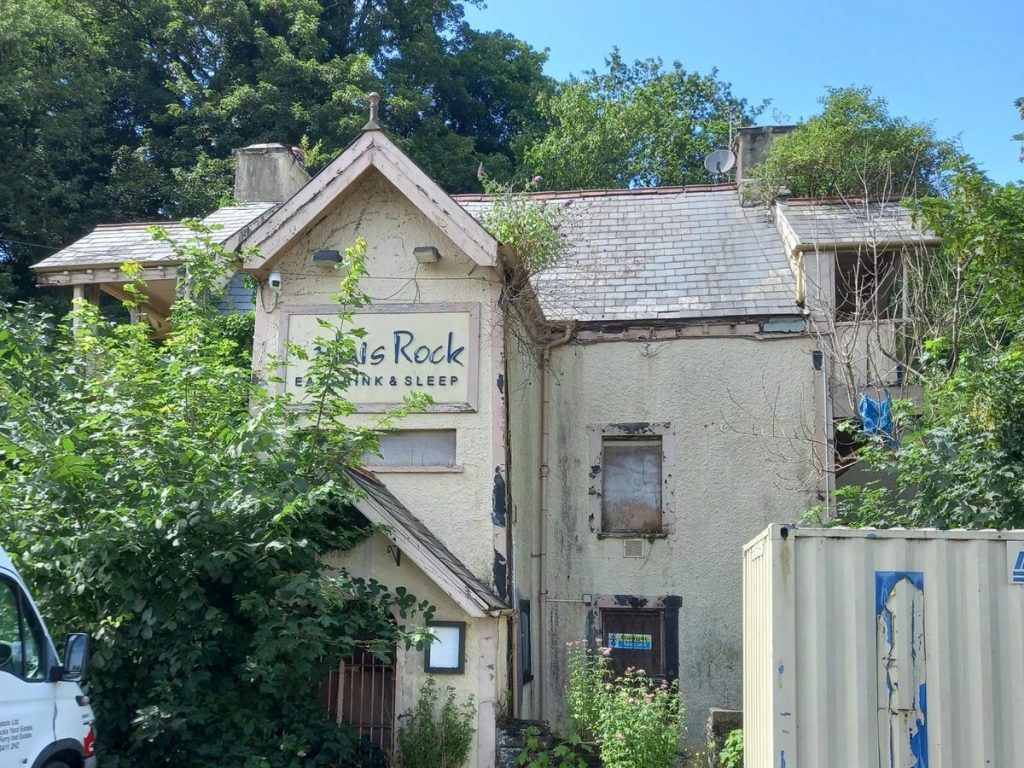 A derelict cream-painted former pub with boarded windows, overgrown with shrubs, a storage container in its car park.