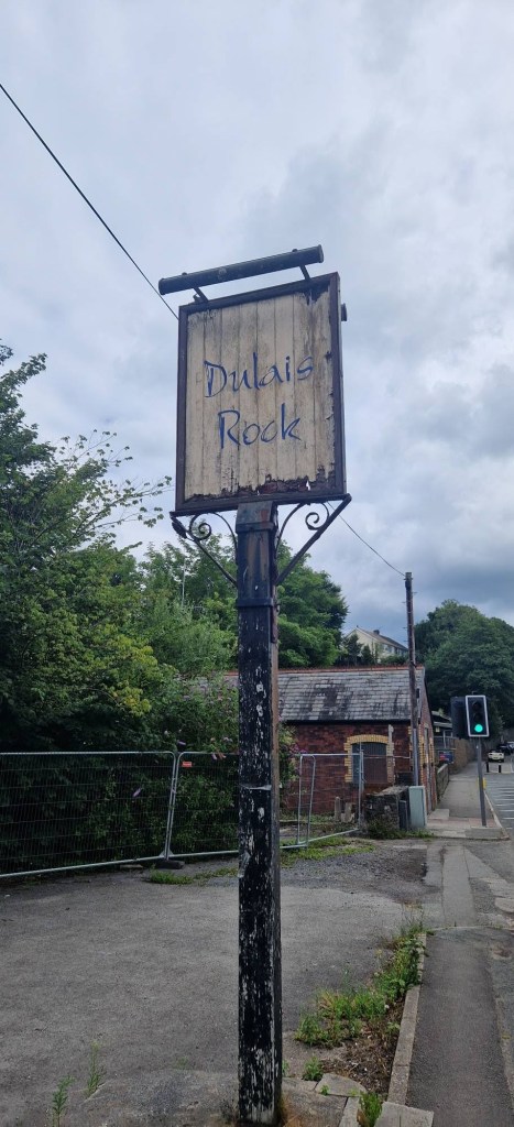 A weathered pub sign reading Dulais Rock on a peeling black post, with security fencing and the boarded pub behind.