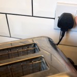 A hand reaching for the switch on a UK plug socket, with a black three-pin plug plugged in next to a toaster