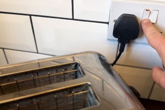 A hand reaching for the switch on a UK plug socket, with a black three-pin plug plugged in next to a toaster