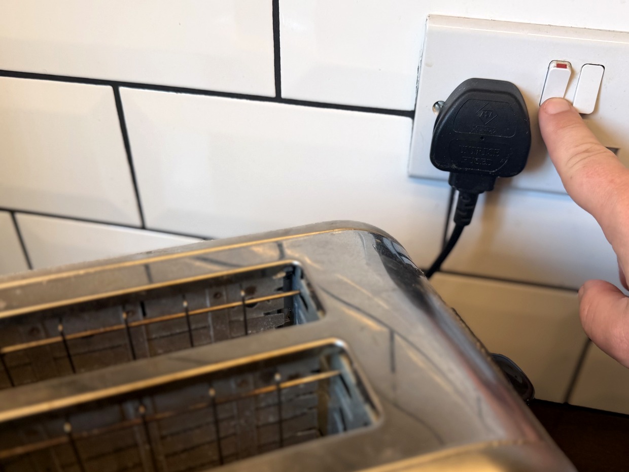 A hand reaching for the switch on a UK plug socket, with a black three-pin plug plugged in next to a toaster