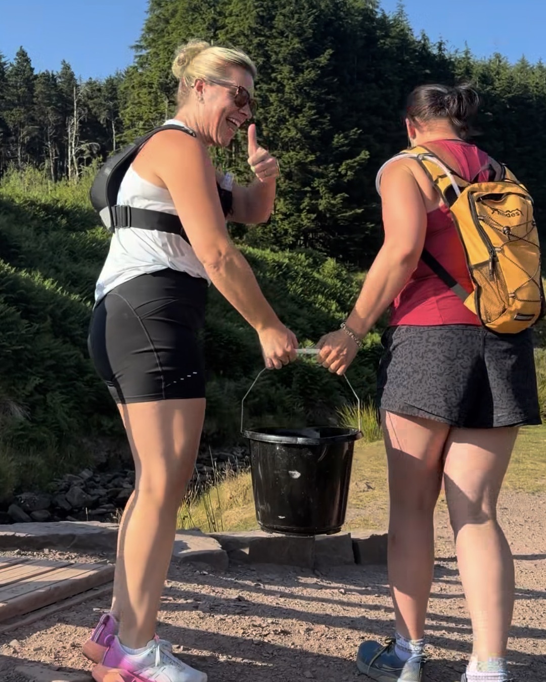 Two women in running gear hand a black bucket between them on a stony path, one giving a thumbs up.