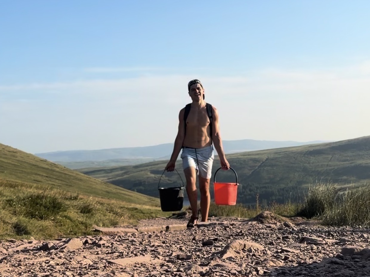 A shirtless man in shorts and sandals carries a black bucket and a red bucket up a steep rocky path, hills and a reservoir behind.