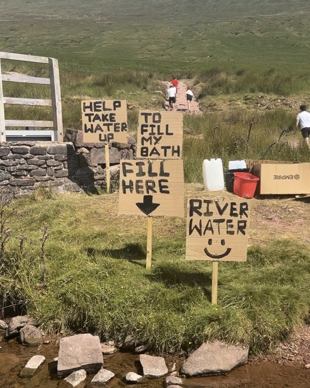 Four hand-lettered cardboard signs staked beside a stream reading Help take water up, To fill my bath, Fill here and River water, with a water container and buckets alongside.