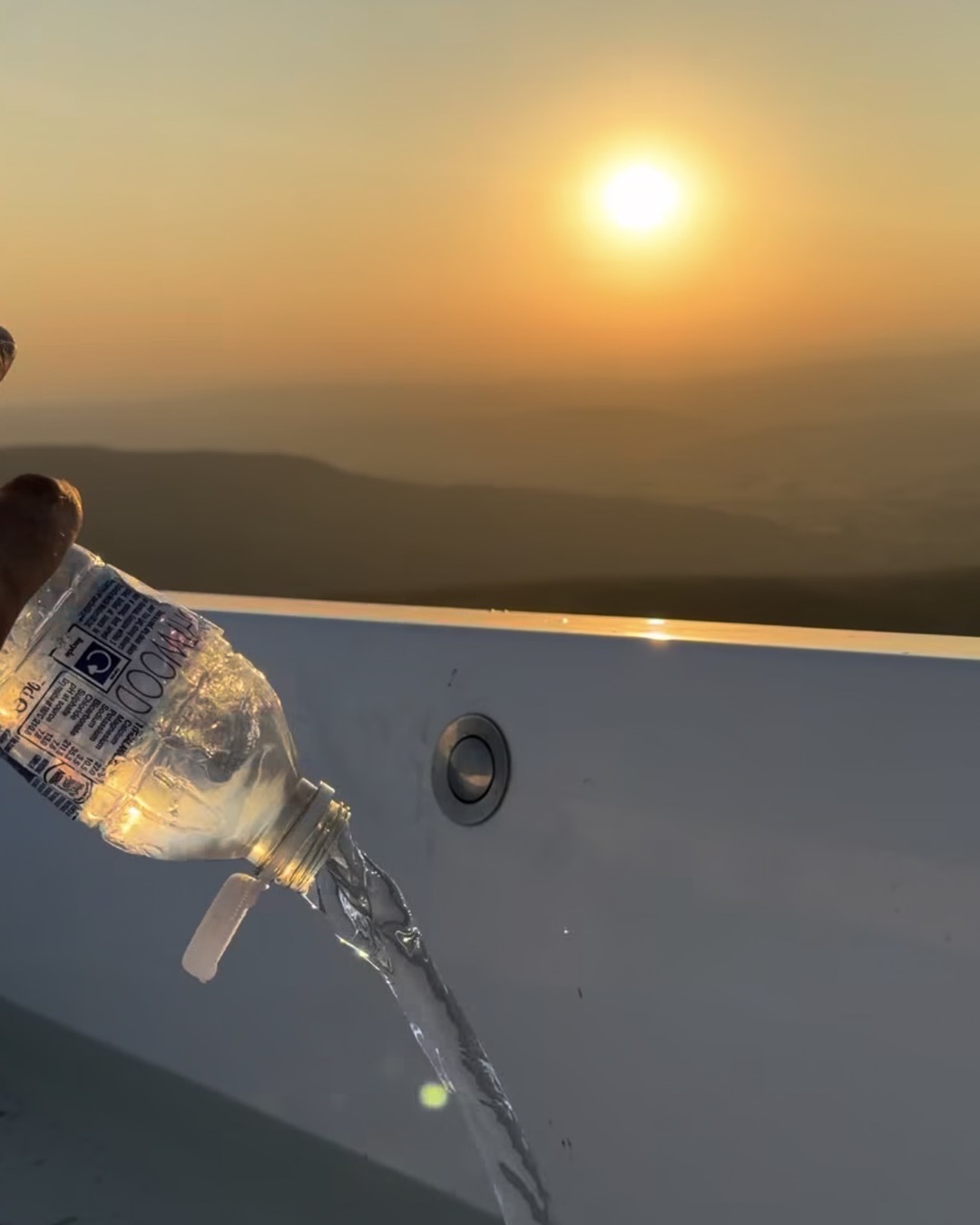 Water pours from a plastic bottle into a white bathtub, the sun low over hazy hills.
