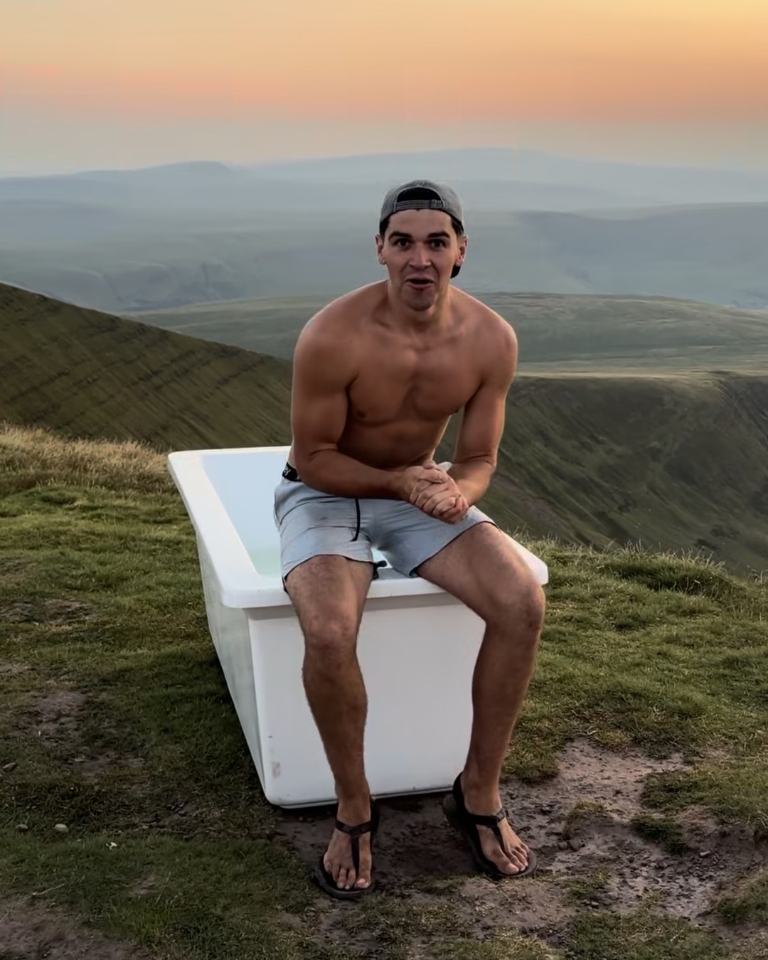 A shirtless man in shorts and sandals sits on the edge of a white bathtub on a mountain summit at sunset, hills fading into haze behind.