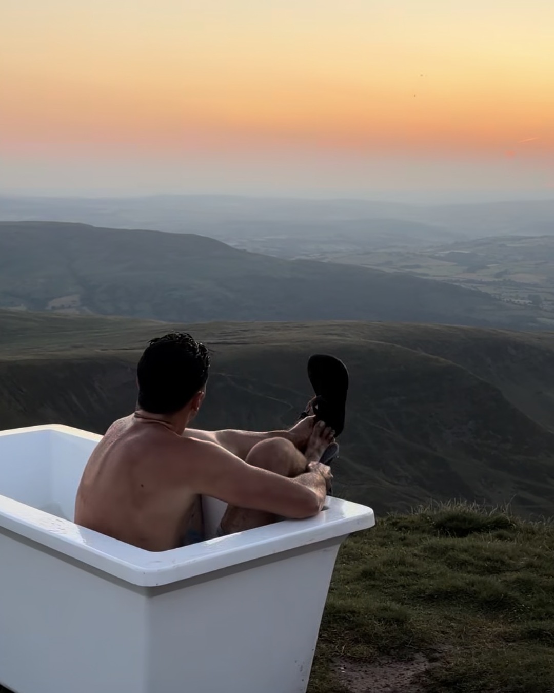 A shirtless man reclines in a white bathtub on a mountain summit at sunset, seen from behind, feet up on the rim.