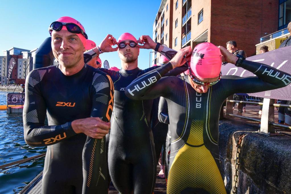 Triathletes in wetsuits and pink swim caps adjust their goggles on the dockside at the Prince of Wales Dock before the start of IRONMAN 70.3 Swansea
