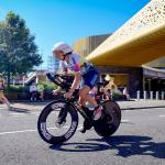 A time-trial cyclist in aero position rides past the copper-clad Swansea Arena during IRONMAN 70.3, with spectators watching from the roadside