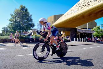 A time-trial cyclist in aero position rides past the copper-clad Swansea Arena during IRONMAN 70.3, with spectators watching from the roadside