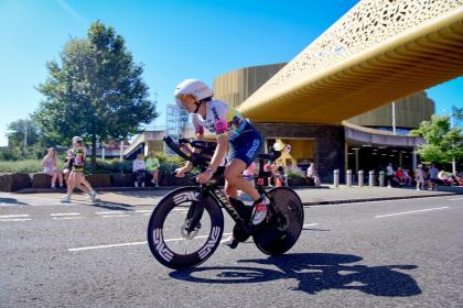 A time-trial cyclist in aero position rides past the copper-clad Swansea Arena during IRONMAN 70.3, with spectators watching from the roadside
