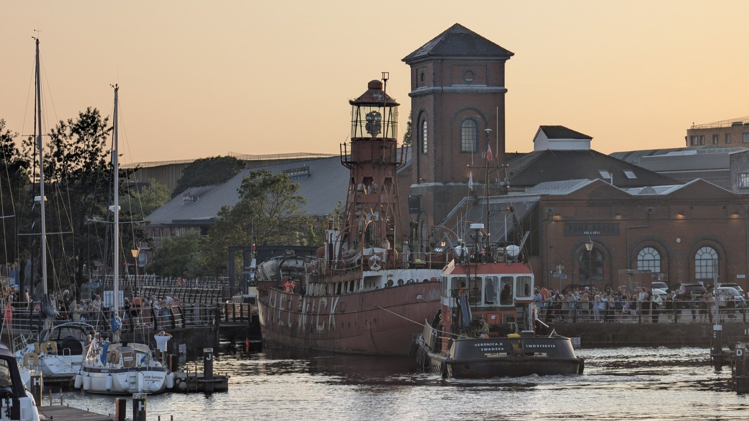 The red Helwick lightship towed by the tug Veronica D past the Pump House in Swansea Marina at sunset, with crowds watching from the quayside