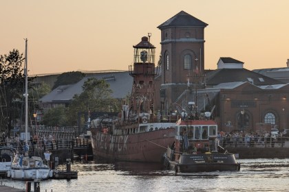 The red Helwick lightship towed by the tug Veronica D past the Pump House in Swansea Marina at sunset, with crowds watching from the quayside