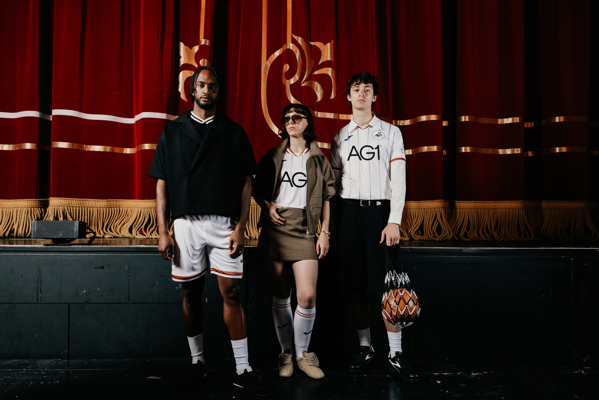 Three models in front of the Grand Theatre's red and gold stage curtain, one wearing the new Swansea City home shirt
