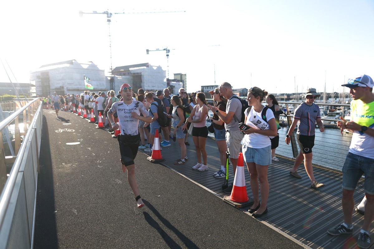 An athlete in a swim cap and goggles runs barefoot across the Sail Bridge lined with spectators