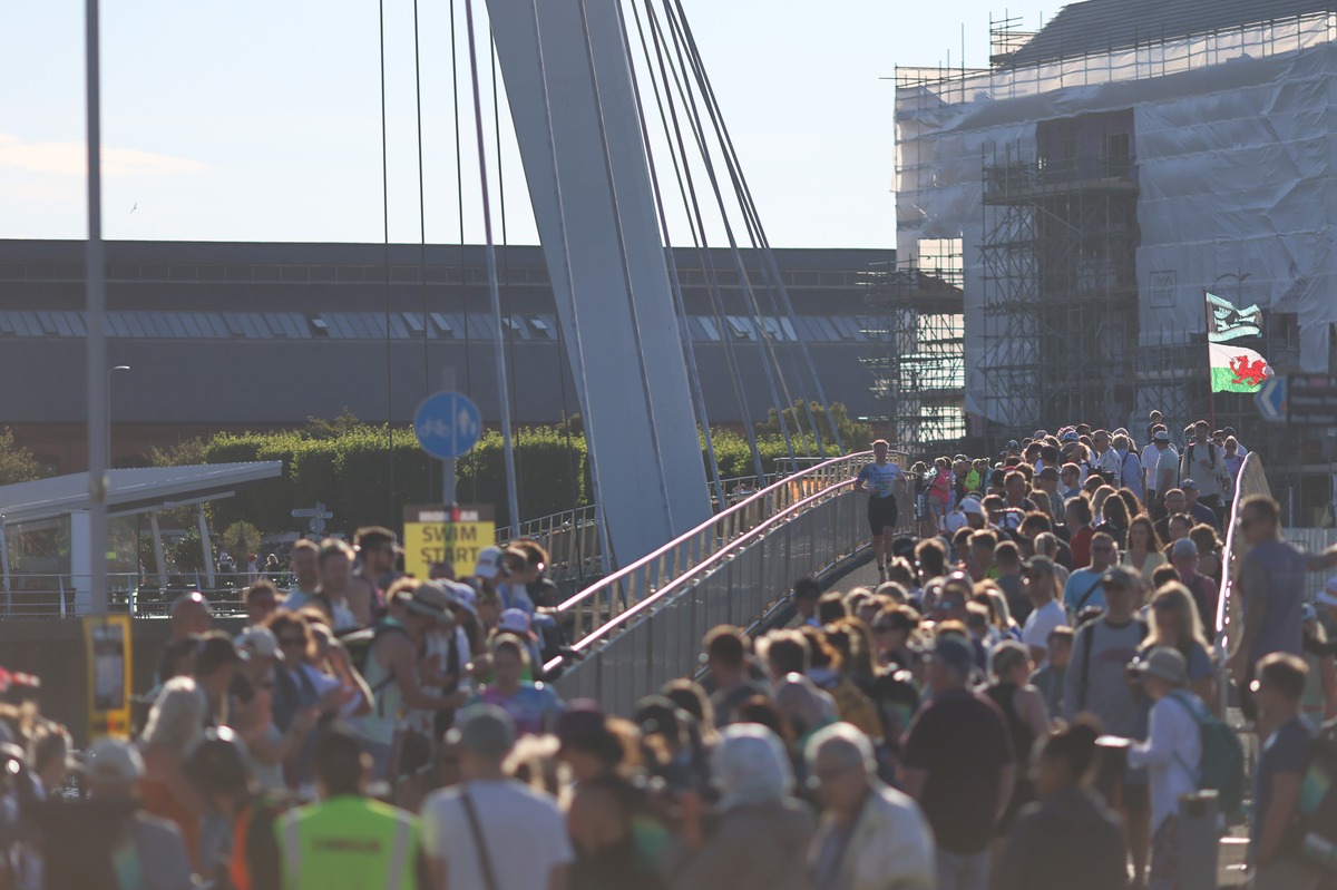Crowds pack the Sail Bridge in morning sunshine with a Welsh flag flying as an athlete runs through