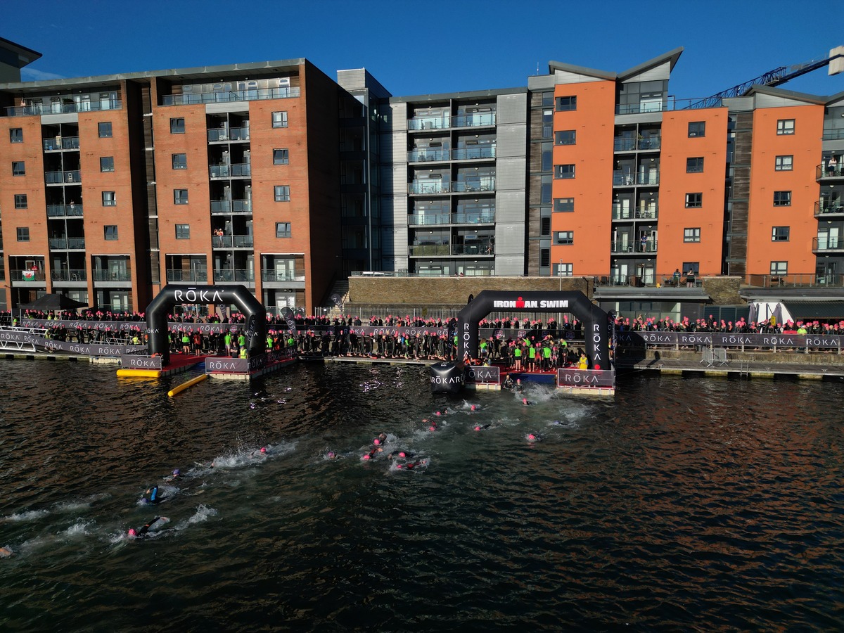 Drone view of swimmers entering the water beneath the IRONMAN swim arch, with a Welsh flag flying from an apartment balcony