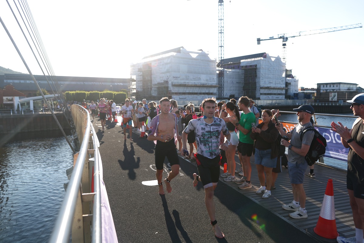 Kieran Lindars runs barefoot from the swim towards transition across the Sail Bridge with another athlete behind