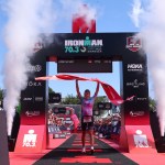 Lizzie Rayner holds the finish tape aloft under the IRONMAN 70.3 Swansea arch, with smoke cannons firing either side