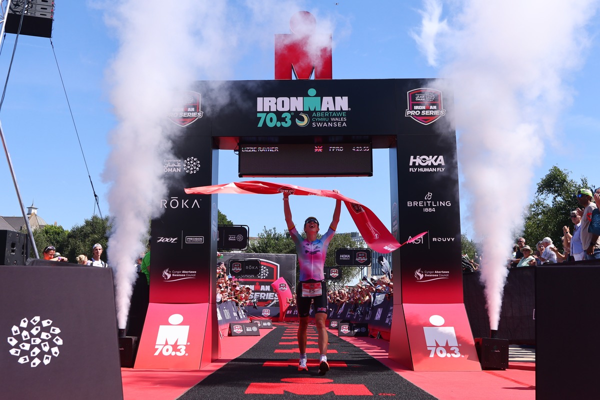Lizzie Rayner holds the finish tape aloft under the IRONMAN 70.3 Swansea arch, with smoke cannons firing either side