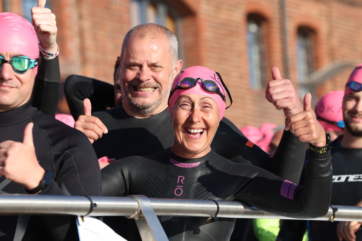 Three smiling athletes in wetsuits and pink swim caps give thumbs up before the start