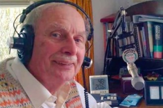 John Powell wearing headphones at a microphone in his home radio studio, smiling, with bookshelves and broadcasting equipment behind him