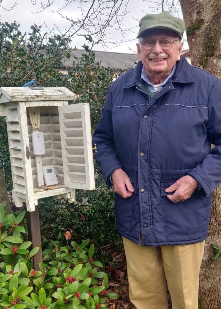 John Powell in a flat cap and navy coat smiling beside an open white louvred weather instrument box in his garden