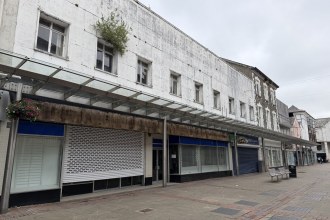 A run of empty shop units with closed roller shutters beneath a weathered glass canopy on Vaughan Street in Llanelli