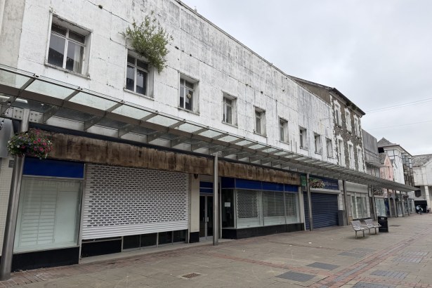 A run of empty shop units with closed roller shutters beneath a weathered glass canopy on Vaughan Street in Llanelli