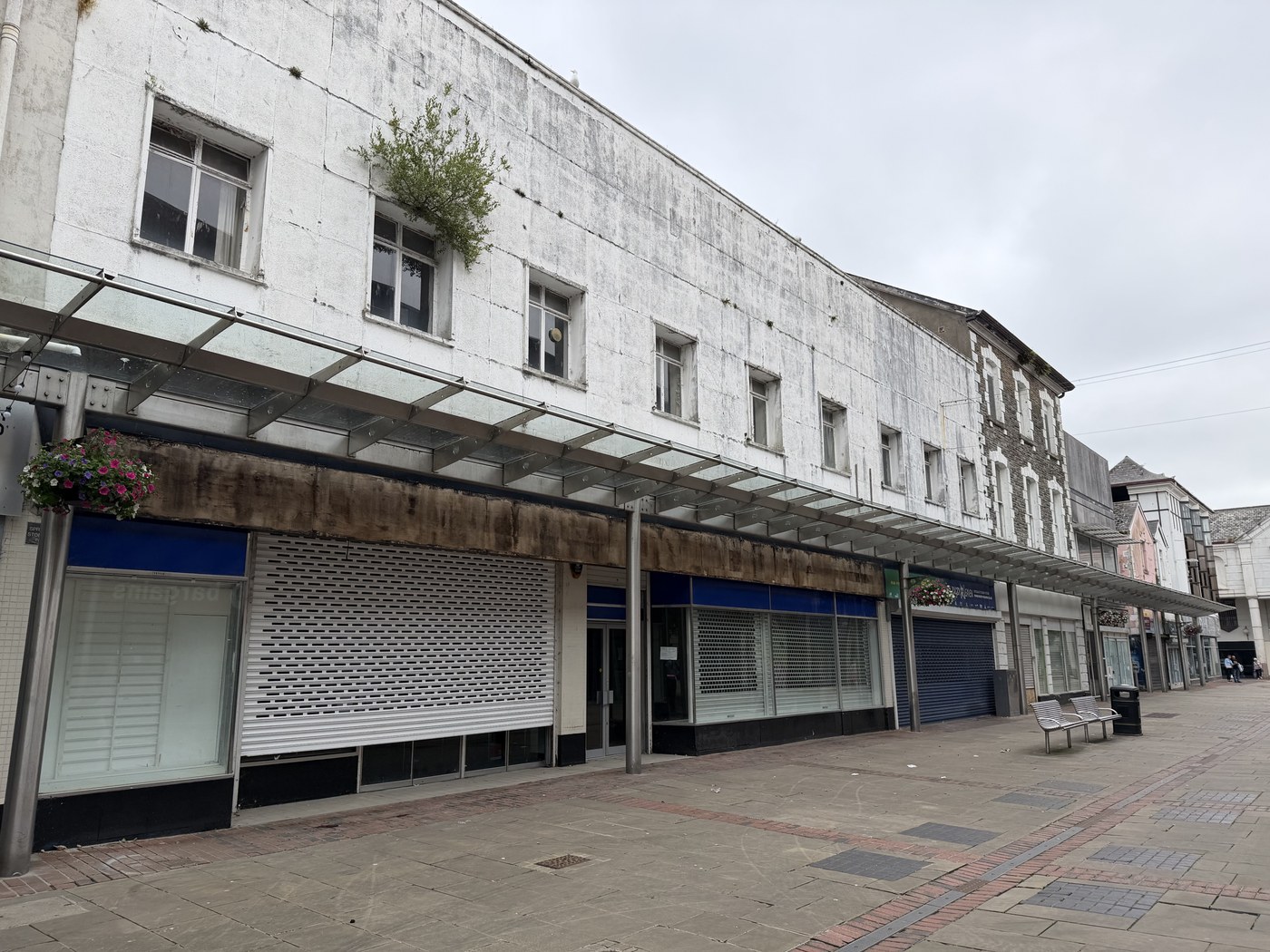 A run of empty shop units with closed roller shutters beneath a weathered glass canopy on Vaughan Street in Llanelli
